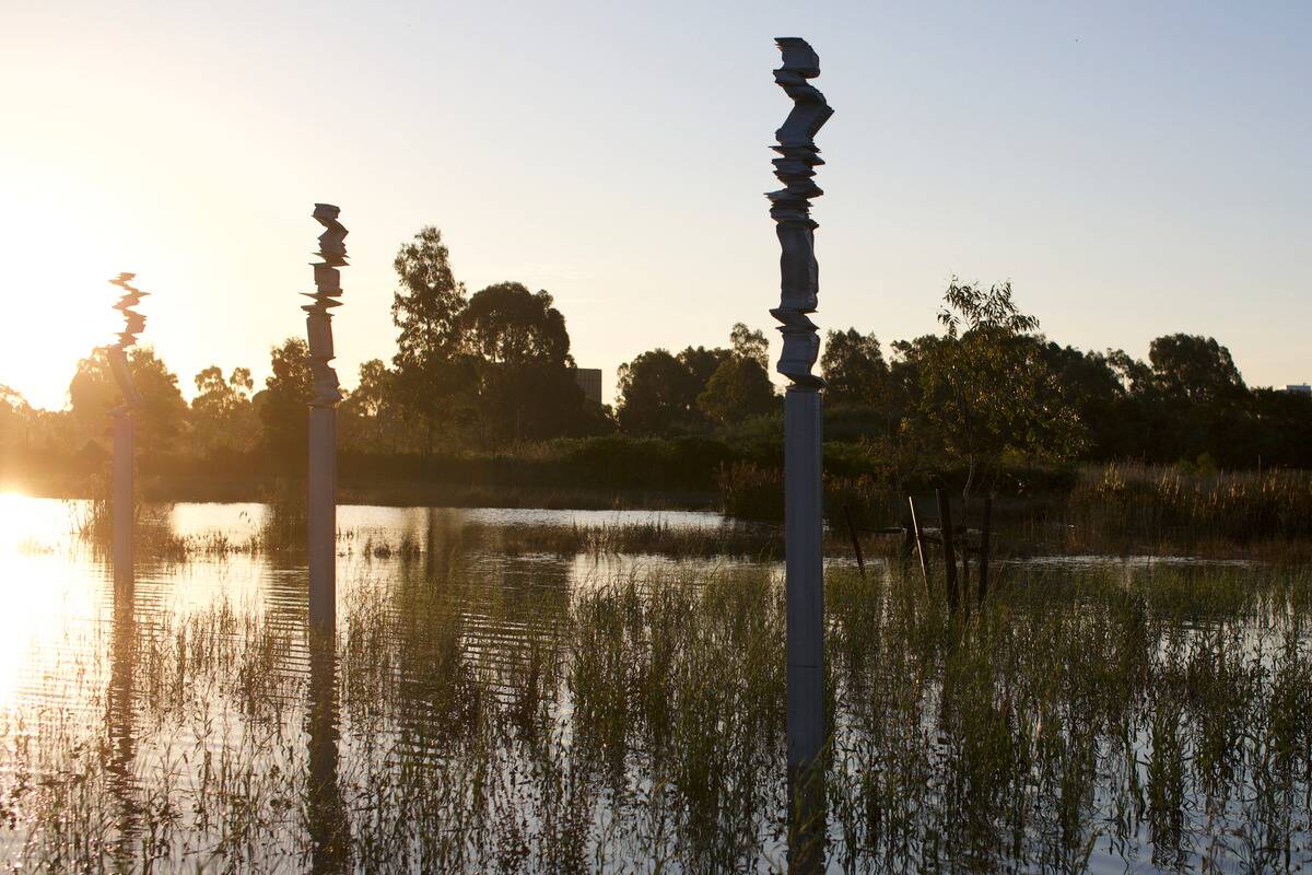 A tranquil landscape at sunset with reflective water partially flooding the area. Four tall, stacked rock sculptures on poles rise from the water. Trees and vegetation are visible in the background, silhouetted against the setting sun.