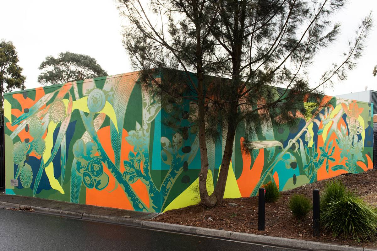 A large, colourful mural featuring various green, orange, and blue abstract shapes and plant-like designs covers the walls of a building. A tree partially obscures the mural. The scene is bordered by a road and some greenery.