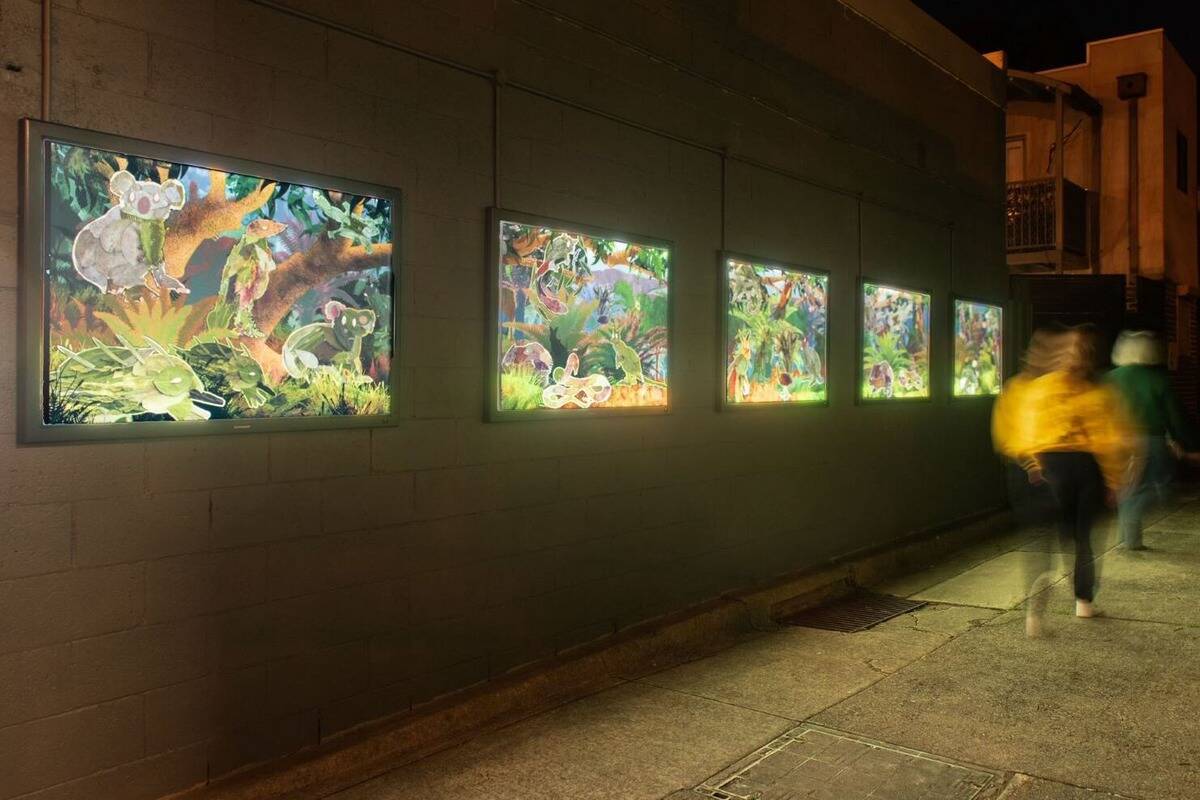 A dimly lit outdoor art exhibition features five illuminated frames displaying vibrant forest scenes. Two blurred figures, one in a yellow jacket and another in darker clothing, walk past the artwork on a concrete pathway. The scene is set at night.