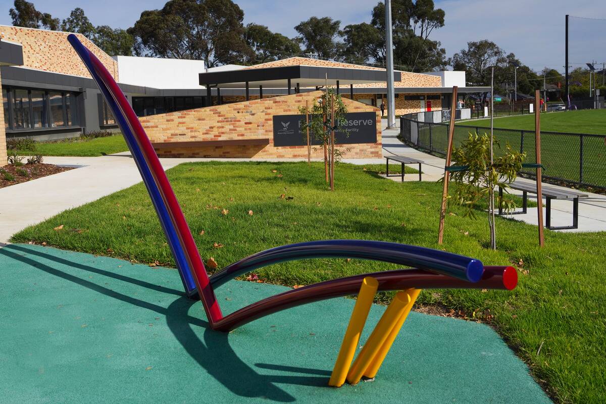 A modern, colourful bench with curved lines sits on a green and blue surface in a well-maintained park. Behind the bench, there are neat grass, trees, a building with brickwork, and a sign reading "Reserve" near a sports field enclosed by a fence.