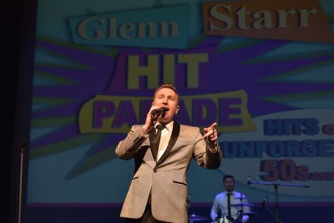 Performer on stage with microphone and wearing beige suit jacket and brown tie singing songs from the 1950's and 1960's in front of a man in a white shirt on a drumkit and screen displaying the performers name and the title of the show.