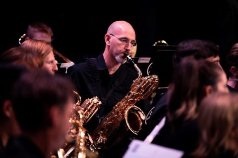 Dark black background, close up of brass section of band, surrounding musicians facing right, slightly at back of heads, focus on mature man with black v-neck top, no hair, short grey beard and glasses blowing into a saxaphone, facing camera with head tilted slightly to the right, looking at his music sheets.