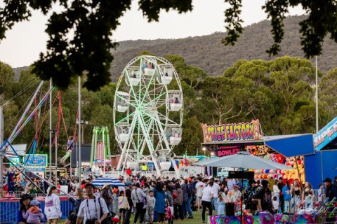 Amusement rides (including ferris wheel, bungee trampoline and carnival games) with a crowd of people walking past, in front of trees and the Dandenong Ranges